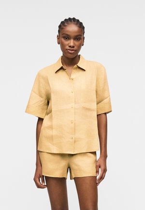 Femme aux cheveux tressés portant une chemise boutonnée jaune clair à manches courtes assortie et un short, debout devant un fond uni.