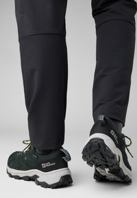 Scarpe outdoor nere con tessuto texture, dettagli verdi e suole in gomma spesse. Indossate con pantaloni scuri, mostrando un primo piano del calzature.