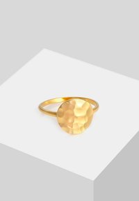 Anello color oro con un design rotondo martellato e una fascia liscia. La superficie riflette la luce, creando texture e profondità variegate.