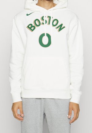 Persoon met een witte hoodie met groene tekst "BOSTON 0" en grijze joggingbroek, staand tegen een witte achtergrond.