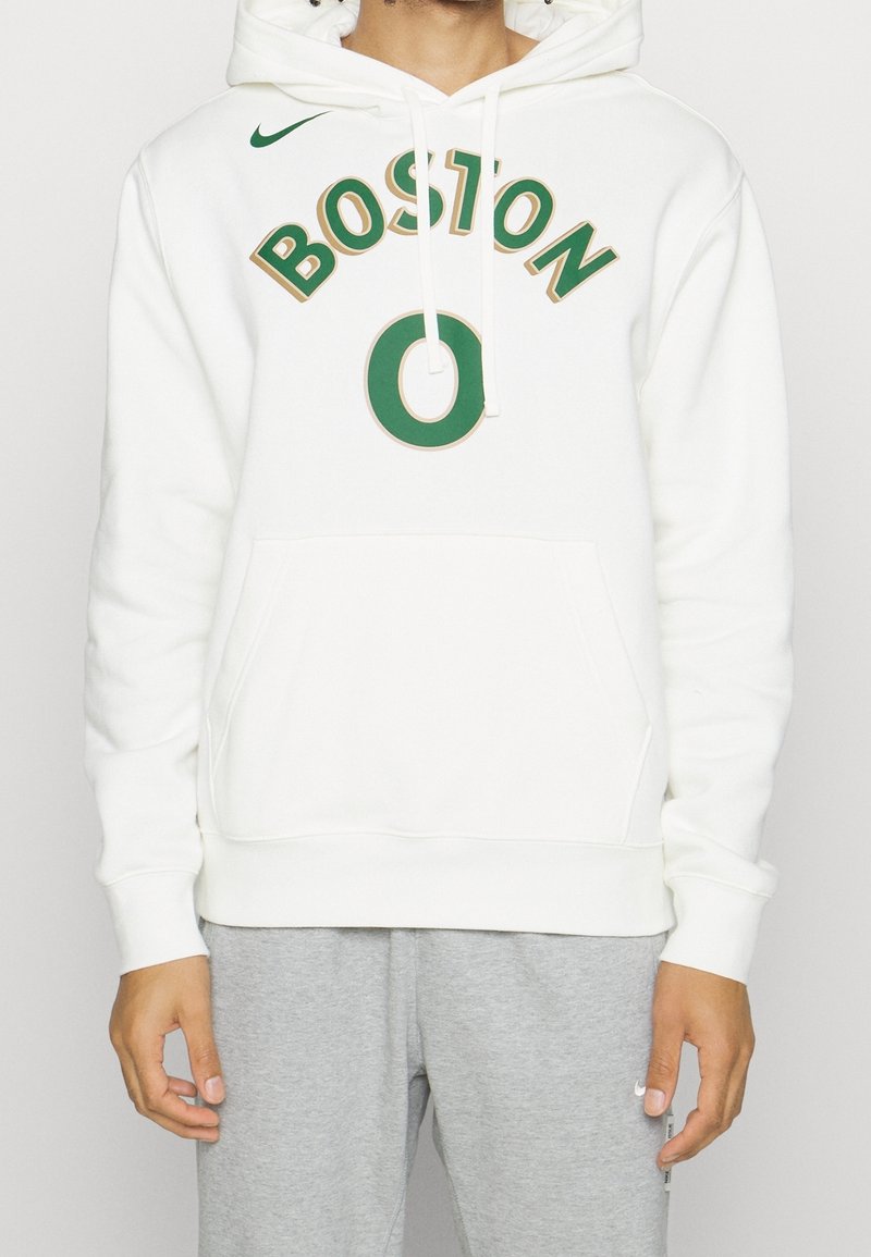 Personne portant un sweat à capuche blanc avec le texte vert « BOSTON 0 » et un pantalon de survêtement gris, se tenant devant un fond blanc.