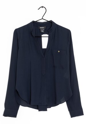 Blouse bleu marine foncé à manches longues avec col mandarin, poche poitrine avec bouton et étiquette, suspendue sur un cintre noir sur fond blanc.