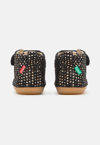 Bottines en daim noir avec des motifs d'étoiles dorées, présentant une semelle en caoutchouc beige et des sangles Velcro. Étiquettes de marque en rouge et vert sur les côtés.