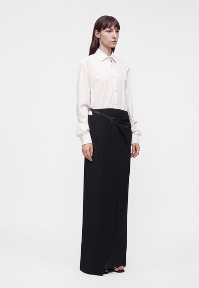 Femme aux longs cheveux bruns portant une chemise blanche boutonnée et une longue jupe portefeuille noire avec des chaussures à bouts ouverts, debout devant un fond uni.
