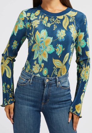 Femme portant un haut à manches longues bleu côtelé avec un motif floral jaune et turquoise, associé à un jean bleu.