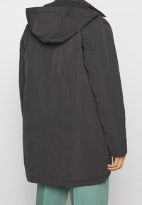 Giacca esterna nera con cappuccio; tessuto leggero e leggermente strutturato; presenta una vestibilità dritta e maniche lunghe, mostrando la silhouette dal retro.
