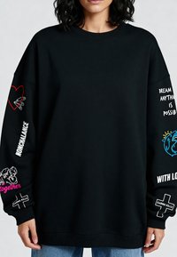 Čierna oversized mikina s grafickými vzormi na rukávoch, vrátane červeného srdca, bieleho textu a modrých akcentov. Rebrované manžety a golier.