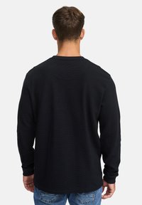 Zwarte blouse met lange mouwen en ribbelstructuur, ronde halslijn en strakke manchetten, gedragen met blauwe jeans. De achterkant toont een rechte zoom.
