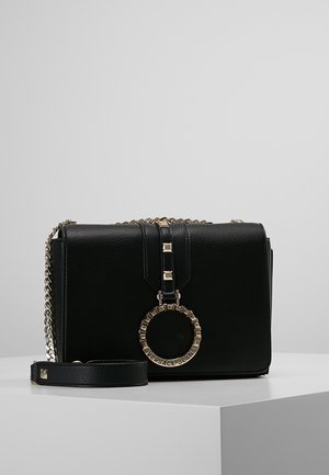 Sac bandoulière en cuir noir avec chaîne dorée et emblème circulaire Versace Jeans sur le rabat avant, posé sur une surface blanche.