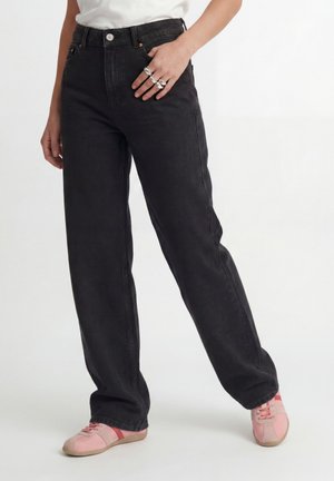 Personne portant un jean noir ample taille haute, un haut blanc, des bagues argentées à la main et des baskets roses à lacets, debout sur fond blanc.