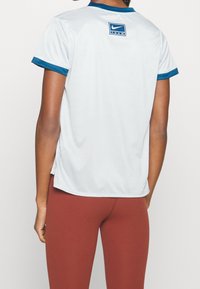 Person som bär en vit sporttröja med blåa detaljer och logotyp på den övre delen av ryggen, i kombination med rostfärgade leggings, visad bakifrån.