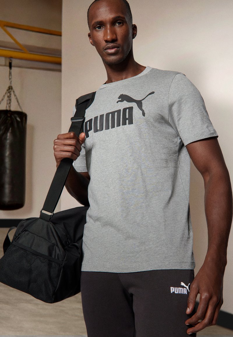 Grå T-skjorte med svart "PUMA"-logo, brukt med svarte treningsbukser. Svart treningsbag har en teksturert bunn og justerbar stropp.