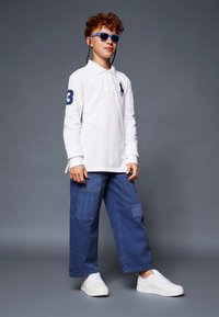 Valge pika varrukaga polo särk tumesinise logo ja numbriga 3, sinised cargo püksid patch-taskutega ja valged tennisetossud. Sinised prillid lõpetavad välimuse.