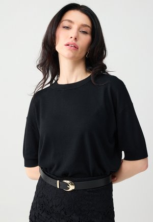 Kobieta z długimi ciemnymi włosami, ubrana w czarny top z krótkim rękawem włożony w czarną koronkową spódnicę z czarnym paskiem z złotą klamrą.