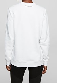 Sudadera blanca de manga larga, con puños y dobladillo acanalados. Presenta la marca "Karl Lagerfeld" en el cuello de la parte trasera en negro. Textura suave.