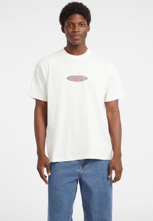 Balts kokvilnas T-krekls ar centrētu ovālu logo, kurā ir redzami "GUESS JEANS" melnā un sarkanā krāsā. Vienkāršs, īsu piedurkņu dizains, brīvas formas.