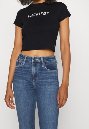 Musta lyhythihainen t-paita, jossa on valkoinen Levi's-logo. Yhdistetty sinisiin korkeavyötäröisiin farkkuihin, joissa on klassinen viiden taskun muotoilu ja etunapit.