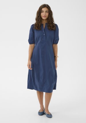 Kobieta z kręconymi brązowymi włosami, ubrana w granatową sukienkę do kolan z bufkami na rękawach i niebieskie baleriny, stojąca na jednolitym tle.