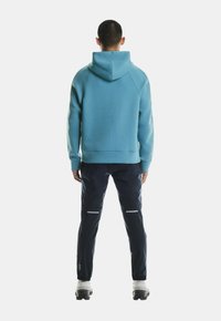 Svetlomodrá mikina s kapucňou a rebrovanými manžetami, skombinovaná s námorníckymi teplákmi s reflexnými detailmi. Model je zobrazený zozadu.