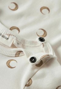 Hvit stoffbody med lysebrune halvmånemønstre. Har svarte trykknapper i halsen og på skulderen for enkel påkledning.