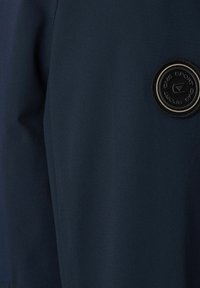 Dunkelblauem Stoff mit gerippter Textur, featuring einem runden schwarzen Patch mit erhabener Schrift und einem Dreiecksdesign.