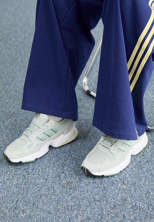 Füße in hellgrauen Turnschuhen und marineblauen Jogginghosen mit gelben Streifen, sitzend auf einem Stuhl auf einem blau getäfelten Teppichboden.