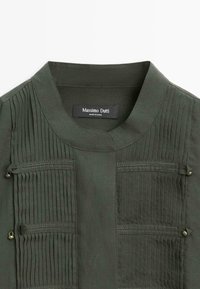 Giacca verde kaki in tessuto morbido con pieghe verticali, collo tondo e tasche con zip arricchite da dettagli decorativi in metallo.