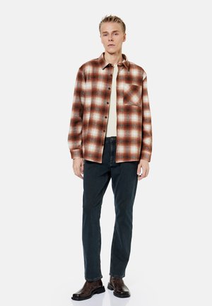 Giovane uomo in piedi che indossa una camicia a quadri marrone e bianca con bottoni, una maglietta beige, jeans neri e stivali marrone scuro su sfondo bianco.