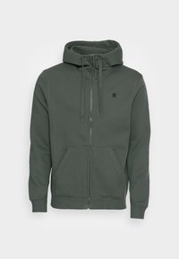 Haut de sweat à capuche zippé vert foncé en tissu doux. Il dispose d'une capuche avec cordon de serrage, de poches avant et d'un petit logo sur la poitrine.