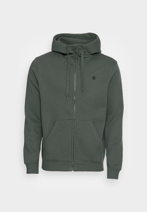 Sudadera con cremallera de color verde oscuro, hecha de un tejido suave. Cuenta con una capucha ajustable, bolsillos frontales y un pequeño logo en el pecho.