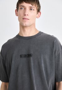 Camiseta de algodón gris con un ajuste relajado. Presenta un cuello redondo y mangas cortas. Destaca un logo negro impreso en el frente. Textura mate.