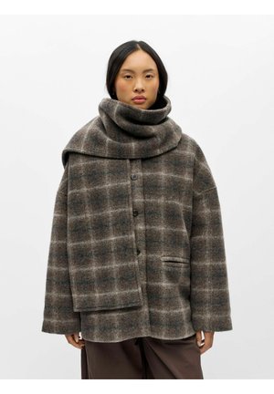 Femme portant un manteau en laine marron et gris à carreaux avec une grande écharpe assortie drapée sur les épaules, sur un fond blanc uni.