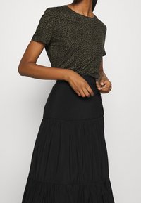 Krátký rukáv olivově zeleného topu s leopardím potiskem, sladěný s černou maxi sukní s volány, která má přiléhavý pas a knoflíkové detaily.