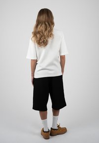 Personne aux longs cheveux ondulés, portant une chemise blanche à manches courtes, un short noir au genou, des chaussettes blanches et des chaussures marron, debout, de dos.