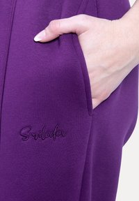 Fialové tepláky s hladkým povrchem, obsahující boční kapsu a vyšitý název značky "Smiledex" blízko lemu.