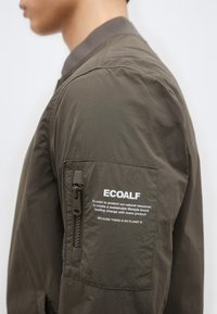 Πλάγια όψη ατόμου που φοράει μπουφάν ελιάς με λογότυπο ECOALF και μήνυμα βιωσιμότητας τυπωμένο στην τσέπη πάνω στο μανίκι.
