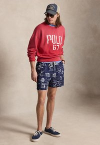 Mladý muž v červené polokošili, tmavě modrých šortkách s květinovým vzorem, modrých plátěných teniskách, slunečních brýlích a tmavě modré a béžové kšiltovce.