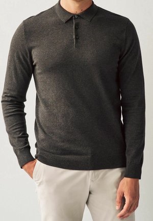 Mężczyzna w ciemnoszarym, długim rękawie polówce z trzema guzikami i jasnobeżowych spodniach, z ręką w kieszeni.