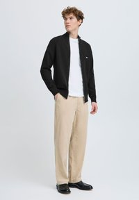 Cardigan nero con zip sopra una maglietta bianca, abbinato a pantaloni beige. Presenta un logo discreto sul cardigan e una texture di tessuto liscia.