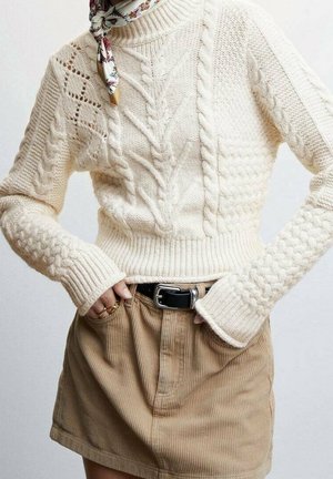 Kobieta ubrana w kremowy, gruby sweter w warkocze, beżową spódnicę sztruksową z czarnym paskiem, kwiecisty szalik na szyi oraz złote pierścionki, z rękami w kieszeniach.