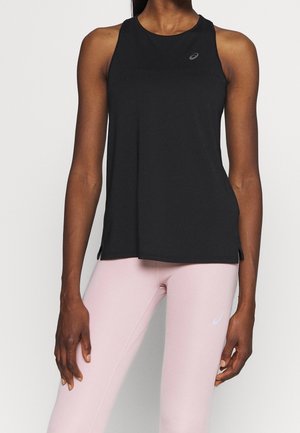 Vrouw die een zwarte mouwloze sporttop en lichtroze legging draagt, staand tegen een effen achtergrond.