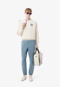 Beige collegepaita, jossa on vihreä brodeerattu logo, siniset verkkarit joustavilla lahkeensuilla, valkoiset lenkkarit ja kermaharmaa laukku mustilla yksityiskohdilla.