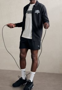 Chaqueta negra con cremallera y logo blanco, camiseta blanca con un logo llamativo, pantalones cortos negros, zapatillas negras y calcetines blancos, sosteniendo una cuerda de saltar.