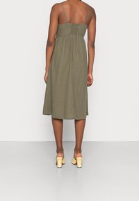 Robe sans manches vert olive avec un corsage ajusté, un dos froncé et une jupe fluide arrivant au milieu du mollet ; portée avec des chaussures à talons blocs jaunes.