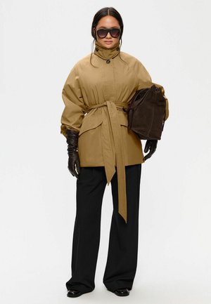 Kvinde iført oversized beige jakke med bælte, sorte vidde bukser, sorte læderhandsker, mørke solbriller, holder stor mørkebrun håndtaske.