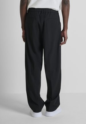 Uomo con pantaloni neri larghi con elastico in vita e sneakers bianche, in piedi contro uno sfondo chiaro e semplice, vista posteriore.