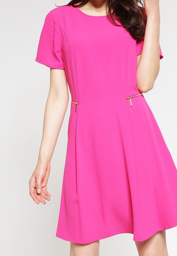 Femme portant une robe rose vif à manches courtes avec deux petites fermetures éclair dorées à la taille, debout devant un fond clair uni.