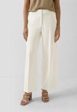 Femme portant un pantalon blanc taille haute à jambes larges et des sandales taupe à talons bloc et bouts ouverts, debout devant un fond gris uni.