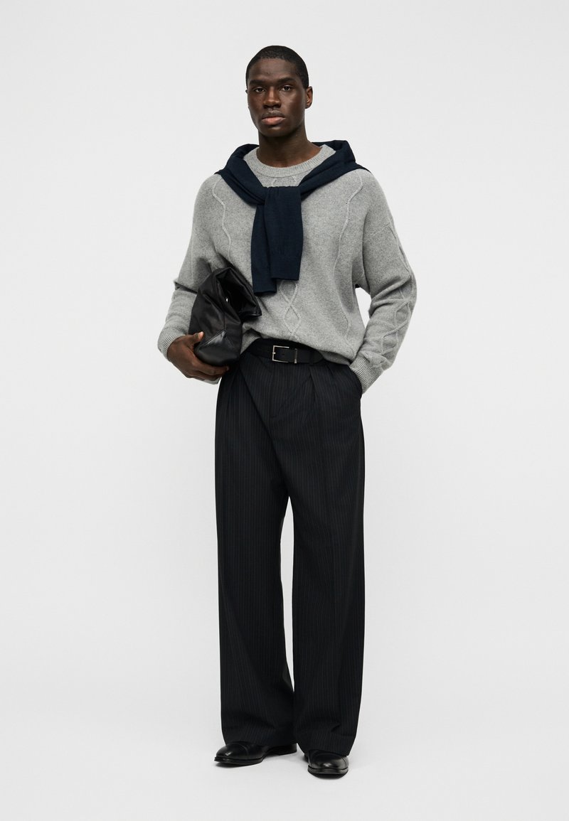 Gray knitted sweater with a diagonal pattern, dark blue scarf draped, dark pinstriped trousers, and black leather shoes held with a black bag.