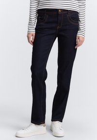 Mørkeblå denimjeans med oransje sting, standard fem-lommer design, rett passform, kombinert med hvite joggesko.